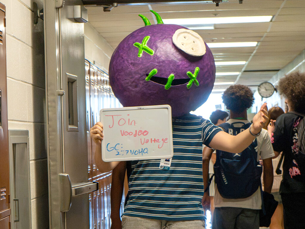 Student with large purple head mask with sign encouraging people to sign up for the robotics team