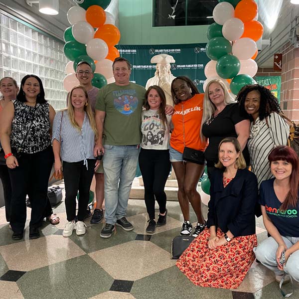 11 people posing for a photo in front of green, white, and orange balloons