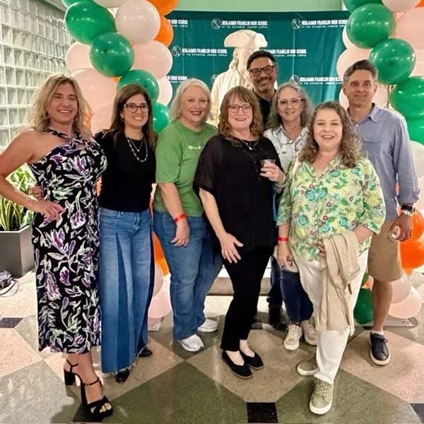 8 adults posing in front of green, white, and orange balloons